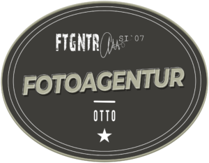 Fotograf Goslar, Logo Fotoagentur Otto, Hochzeitsfotograf. Logo der Fotoagentur Otto: Ein ovales, graues Emblem mit einem dreifachen Rand in unterschiedlichen Grautönen. Im oberen Bereich steht in einer weißen, abgenutzten Schrift 'FTGNTR' und daneben 'SI '07'. In der Mitte befindet sich der Schriftzug 'FOTOAGENTUR' in großen, schattierten Buchstaben. Darunter ist ein horizontaler Strich und darunter steht 'OTTO' in weißer Schrift. Am unteren Rand des Logos ist ein weißer Stern abgebildet. Fotograf aus Goslar