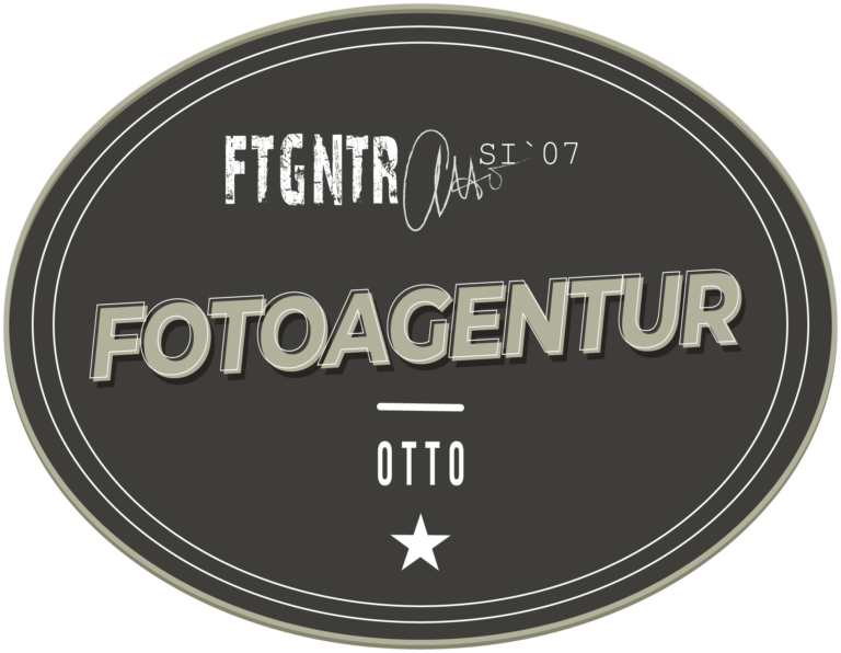 Fotograf Goslar, Logo Fotoagentur Otto, Hochzeitsfotograf. Logo der Fotoagentur Otto: Ein ovales, graues Emblem mit einem dreifachen Rand in unterschiedlichen Grautönen. Im oberen Bereich steht in einer weißen, abgenutzten Schrift 'FTGNTR' und daneben 'SI '07'. In der Mitte befindet sich der Schriftzug 'FOTOAGENTUR' in großen, schattierten Buchstaben. Darunter ist ein horizontaler Strich und darunter steht 'OTTO' in weißer Schrift. Am unteren Rand des Logos ist ein weißer Stern abgebildet. Fotograf aus Goslar