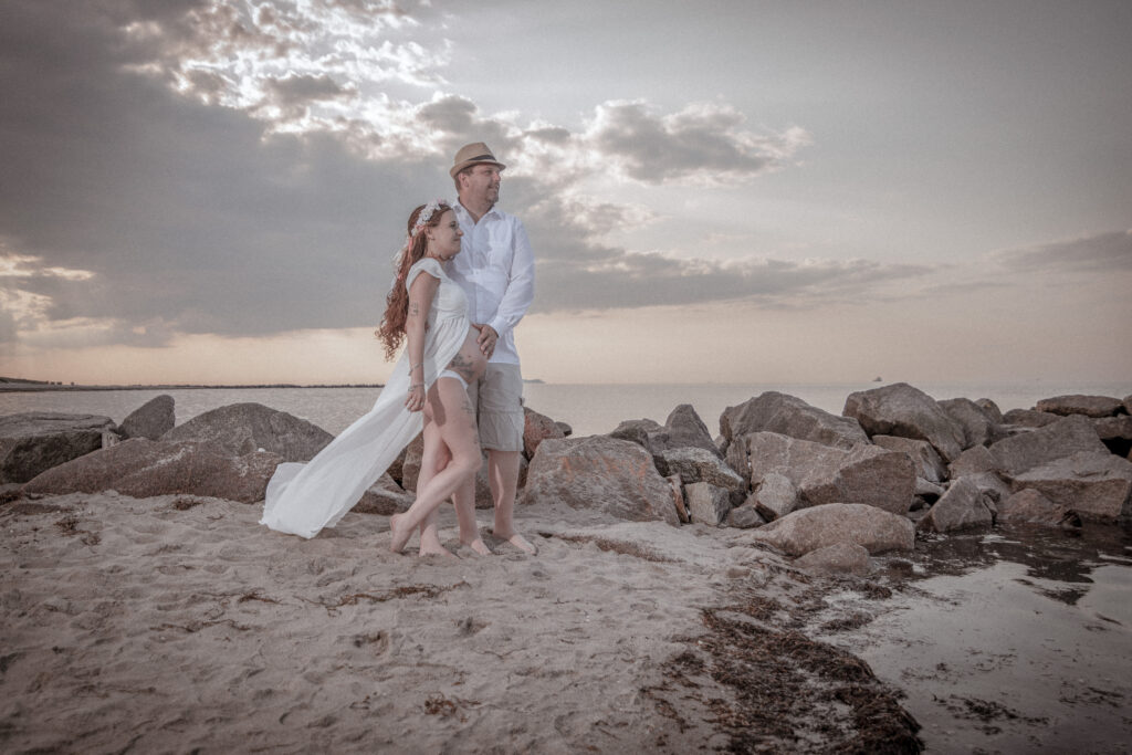 Ein sonniger Strandtag, eine schwangere Braut strahlt in ihrem Hochzeitskleid, während sie am Ufer entlang spaziert