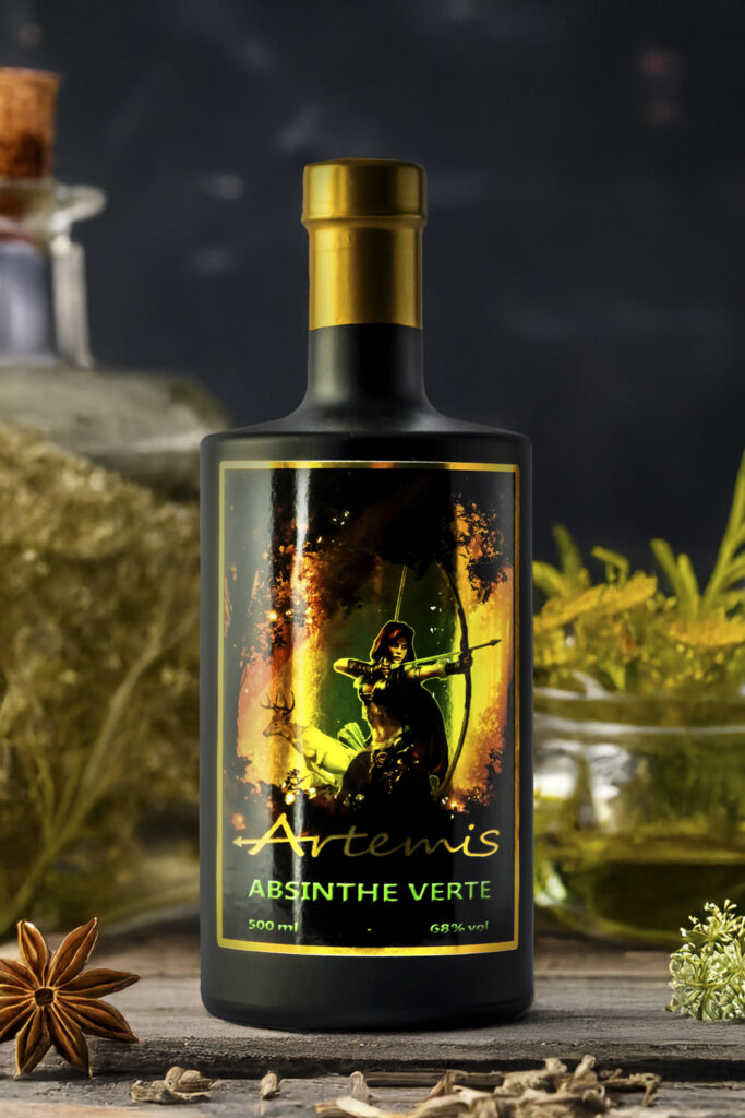 Produktfoto der Fotoagentur Otto aus Goslar – edle schwarze Flasche „Artemis Absinthe Verte“ mit goldenem Verschluss, künstlerischem Etikett und natürlichen Kräutern im Hintergrund. Professionelle Food- und Produktfotografie.