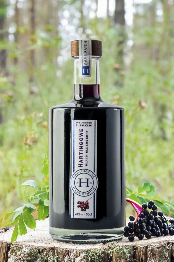 Produktfoto der Fotoagentur Otto aus Goslar – edle Flasche „Hartingowe Holunder Likör“ Black Elderberry, stilvoll platziert auf einem Baumstamm mit frischen Holunderbeeren im Vordergrund, im natürlichen Waldhintergrund fotografiert. Professionelle Produktfotografie.