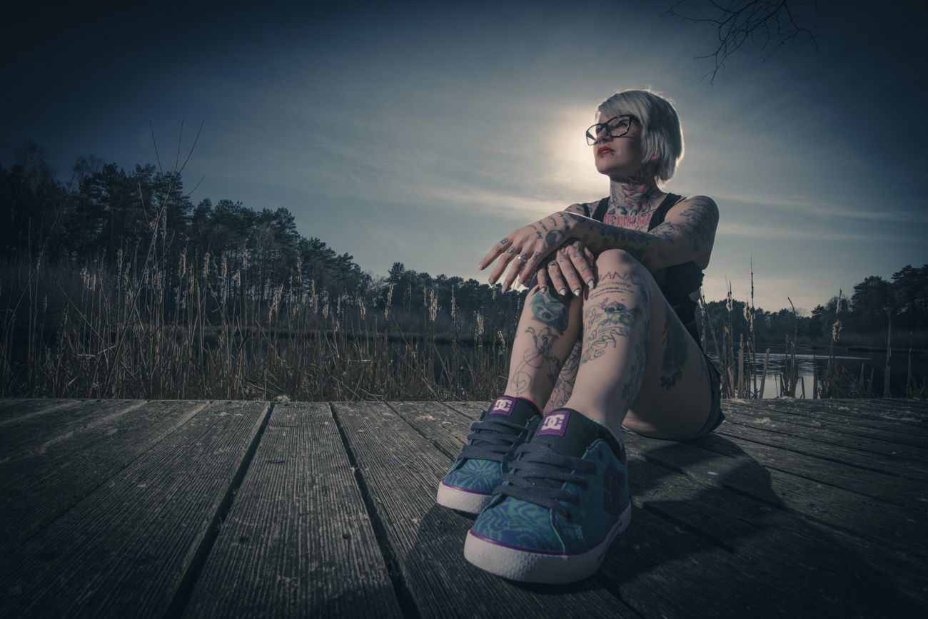 Kreatives Porträt von Fotoagentur Otto in Goslar: Tätowierte Frau mit Brille sitzt auf einem Holzsteg am See und blickt nachdenklich in die Sonne. Ausdrucksstarke und individuelle Porträtfotografie aus Goslar von Fotoagentur Otto.